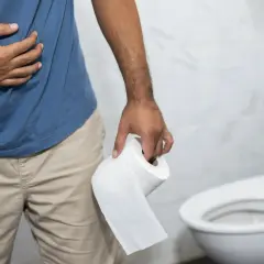 Mężczyzna trzymający papier toaletowy, z ręką na brzuchu, w łazience. Wygląda na to, że ma **biegunkę po alkoholu**.