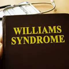 Dłoń trzyma książkę z napisem "Williams Syndrome". Obok leży stetoskop i długopis.